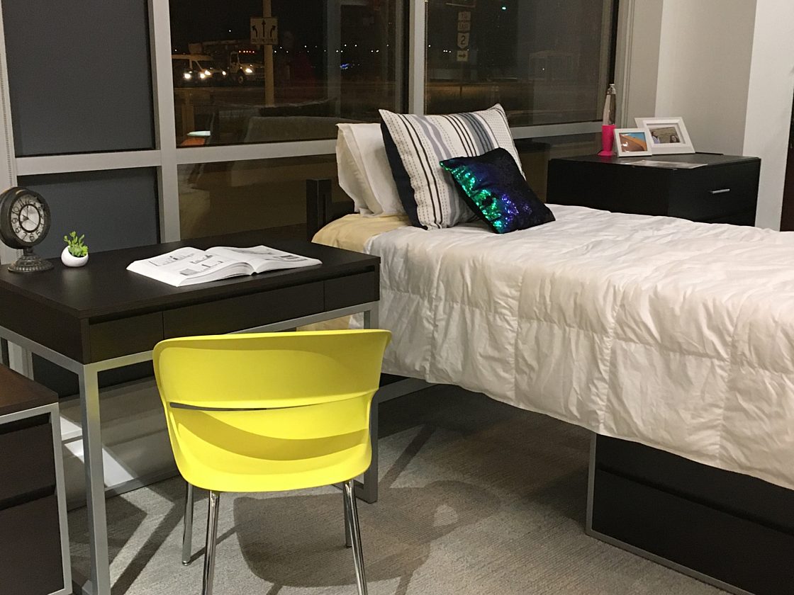 A mock-up of a college dorm bed with a bedside cabinet and a small desk. The space is decorated with colorful pillows and knickknacks.