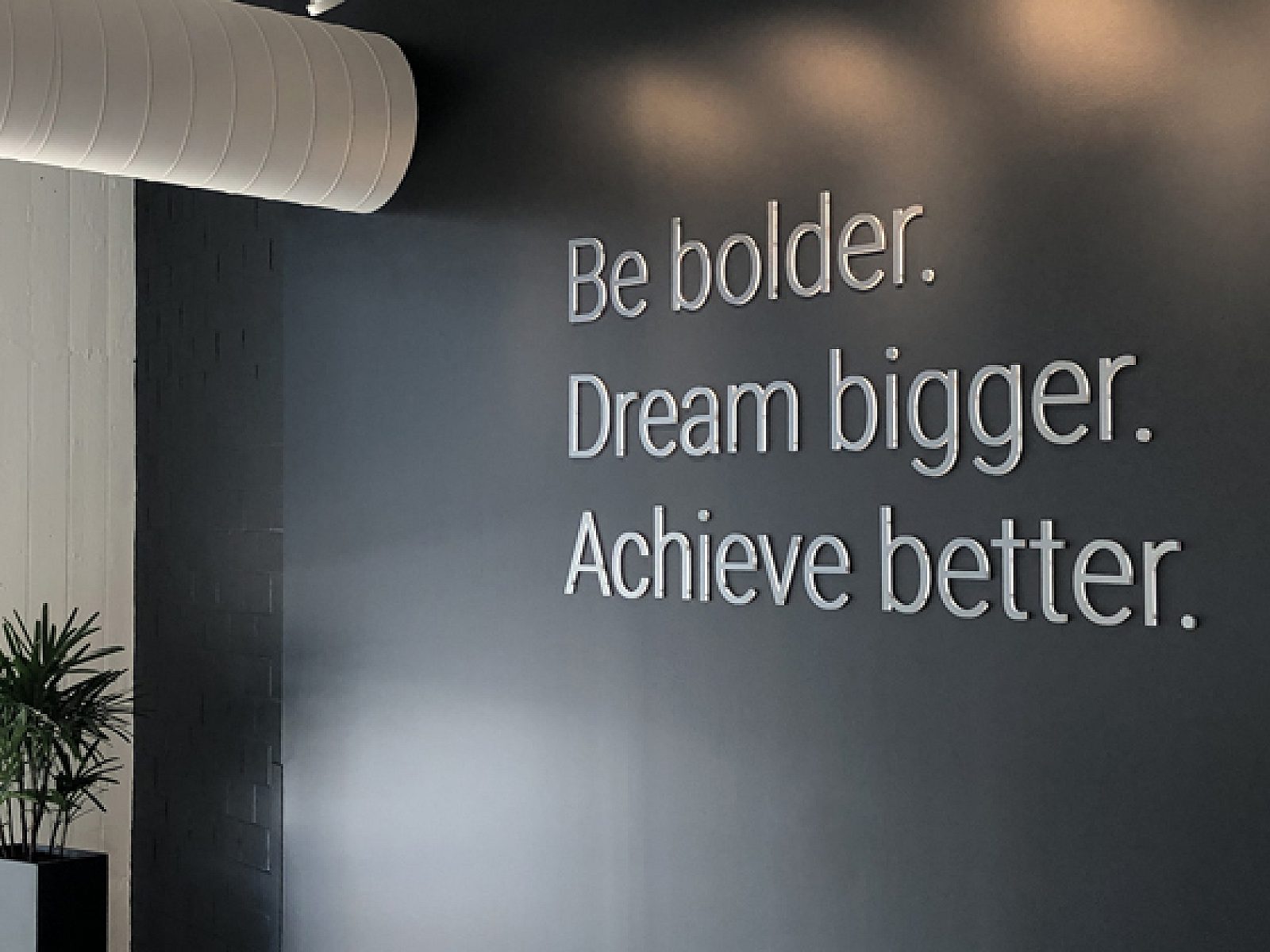 View of dark grey wall with inspirational brand message: Be bolder, dream bigger, achieve better. Windows to the left let in bright sunlight.