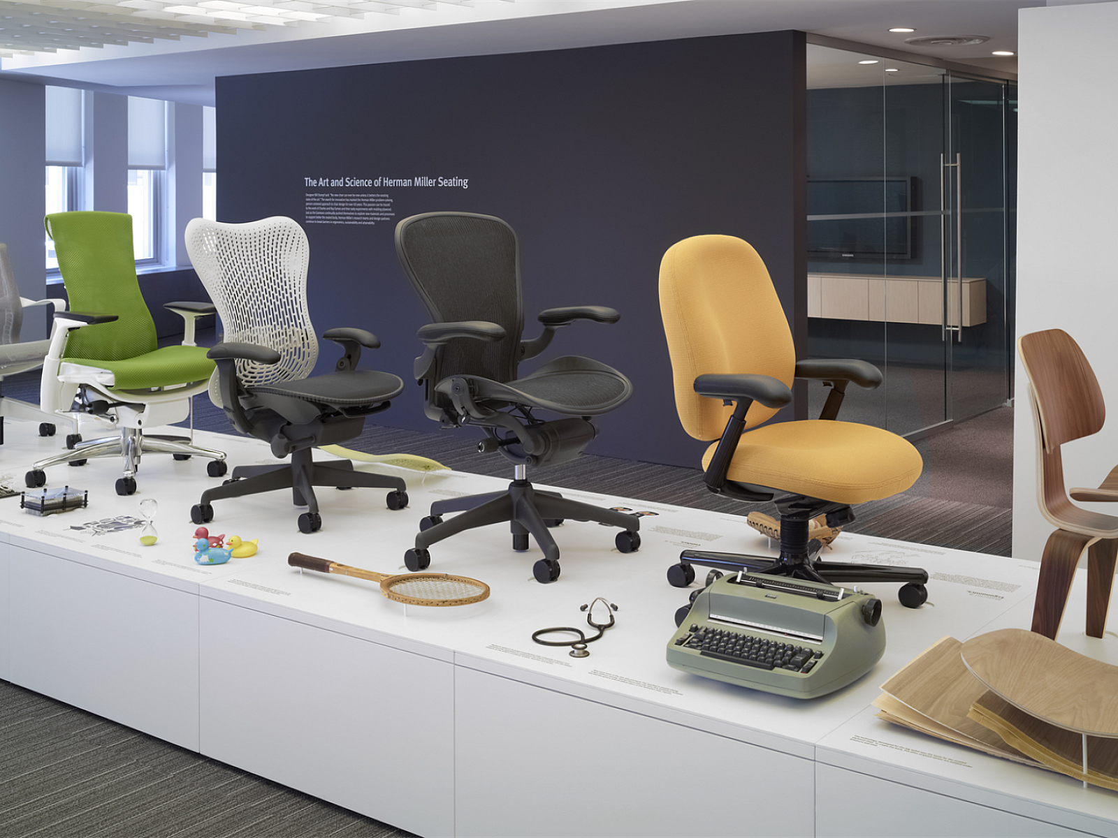 A raised display of Herman Miller chairs of over the years: Eames Molded Plywood, Aeron, Embody, Setu, Sayl, alongside historic Eames leg splint