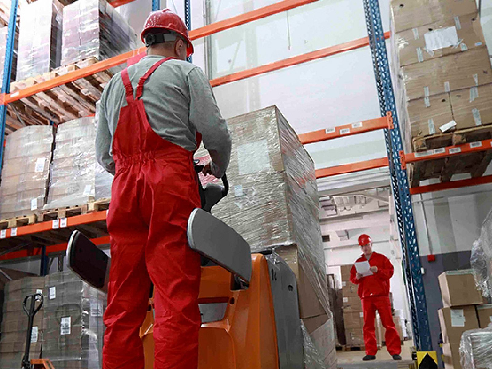 Low-angle view of a forklift operator in a bright warehouse. Tall shelving full of wrapped pallets, supervisor in distance with clipboard.