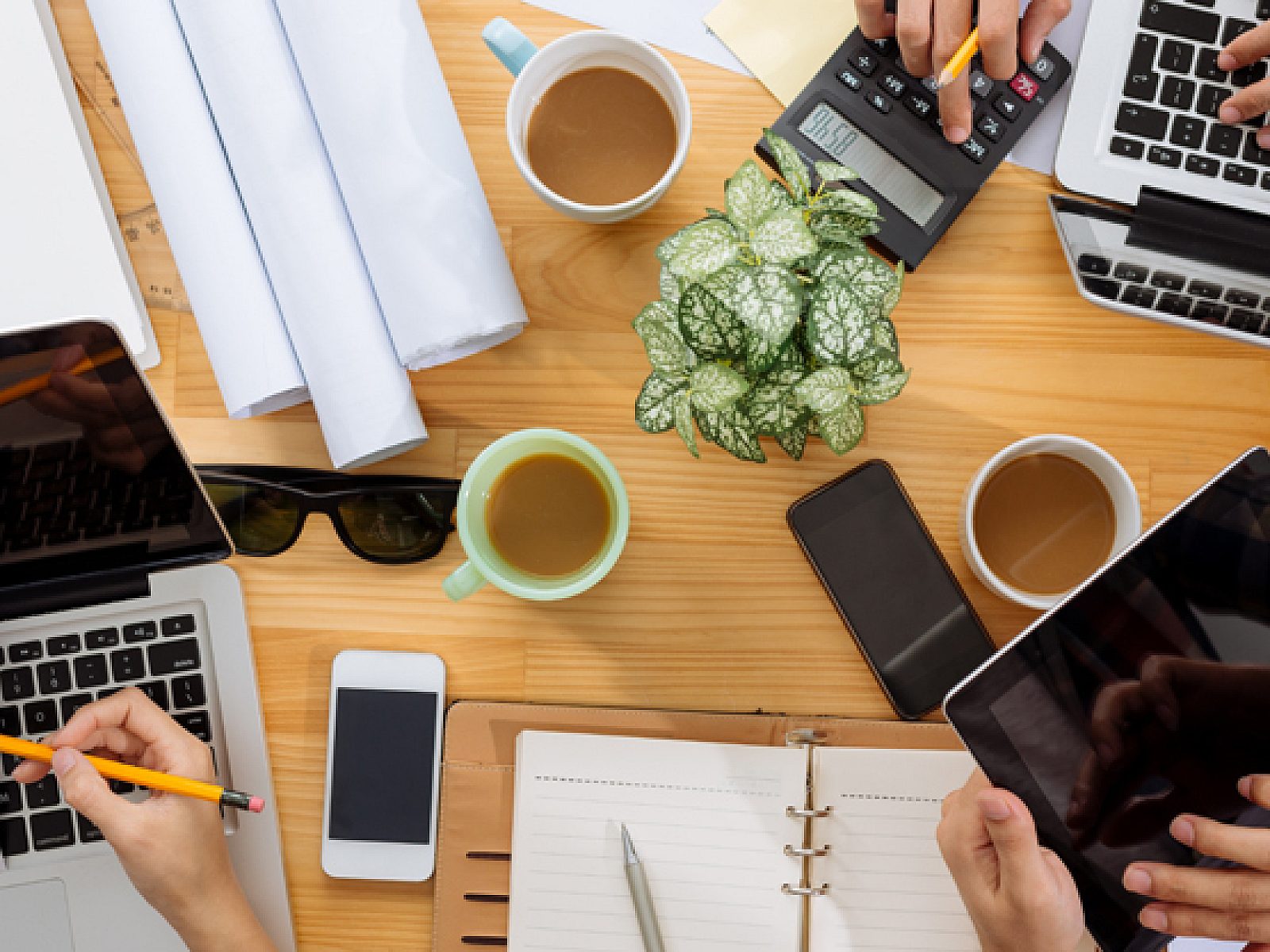 Overhead view of people at a work table: hands, laptops, drawings, planners, pencils, coffee mugs