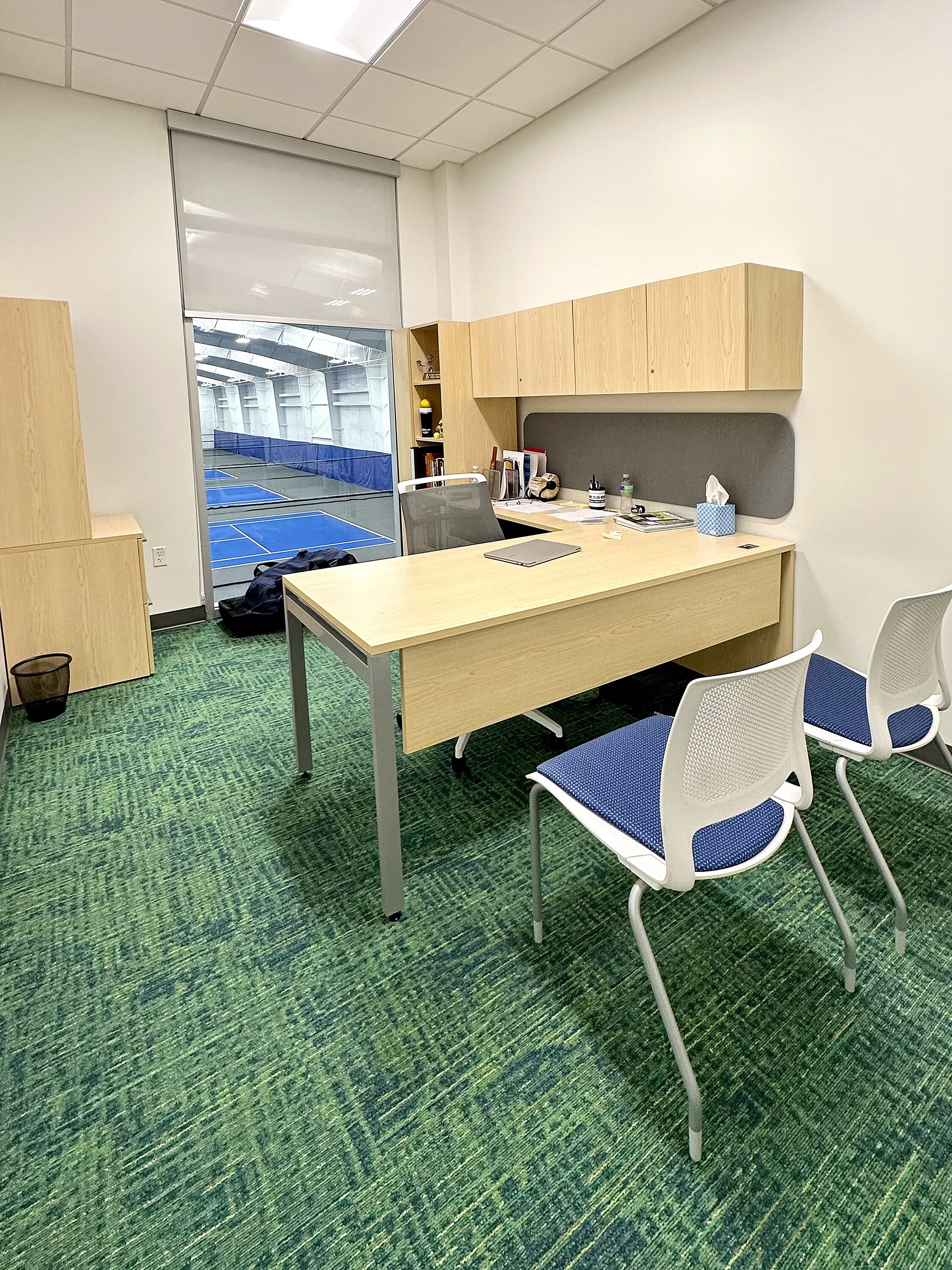 A private office overlooking the indoor tennis courts. Features L-shaped desk with overhead cabinets, plenty of storage, and two guest chairs.