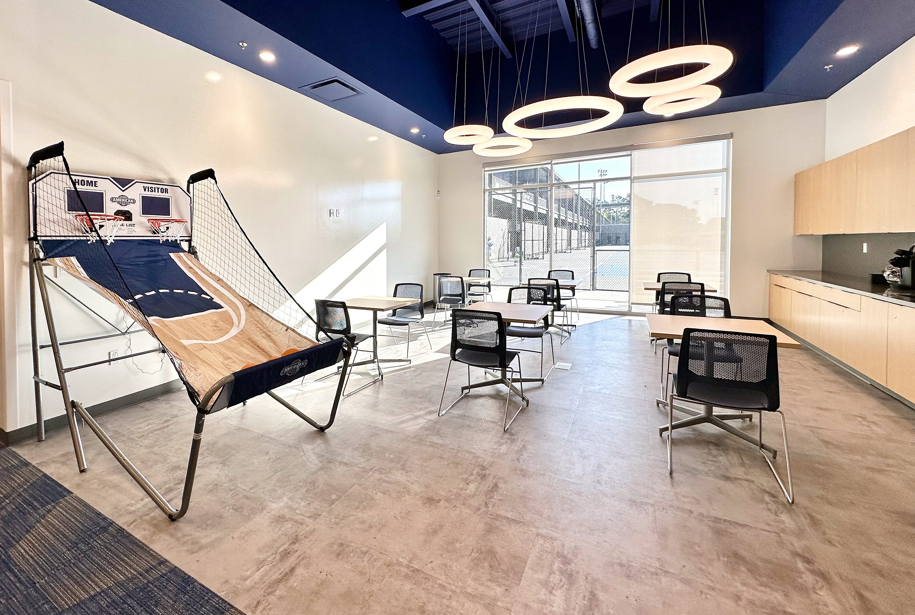 Contemporary café with small tables, circular pendant lights, a basketball arcade game, large windows, and a kitchenette.
