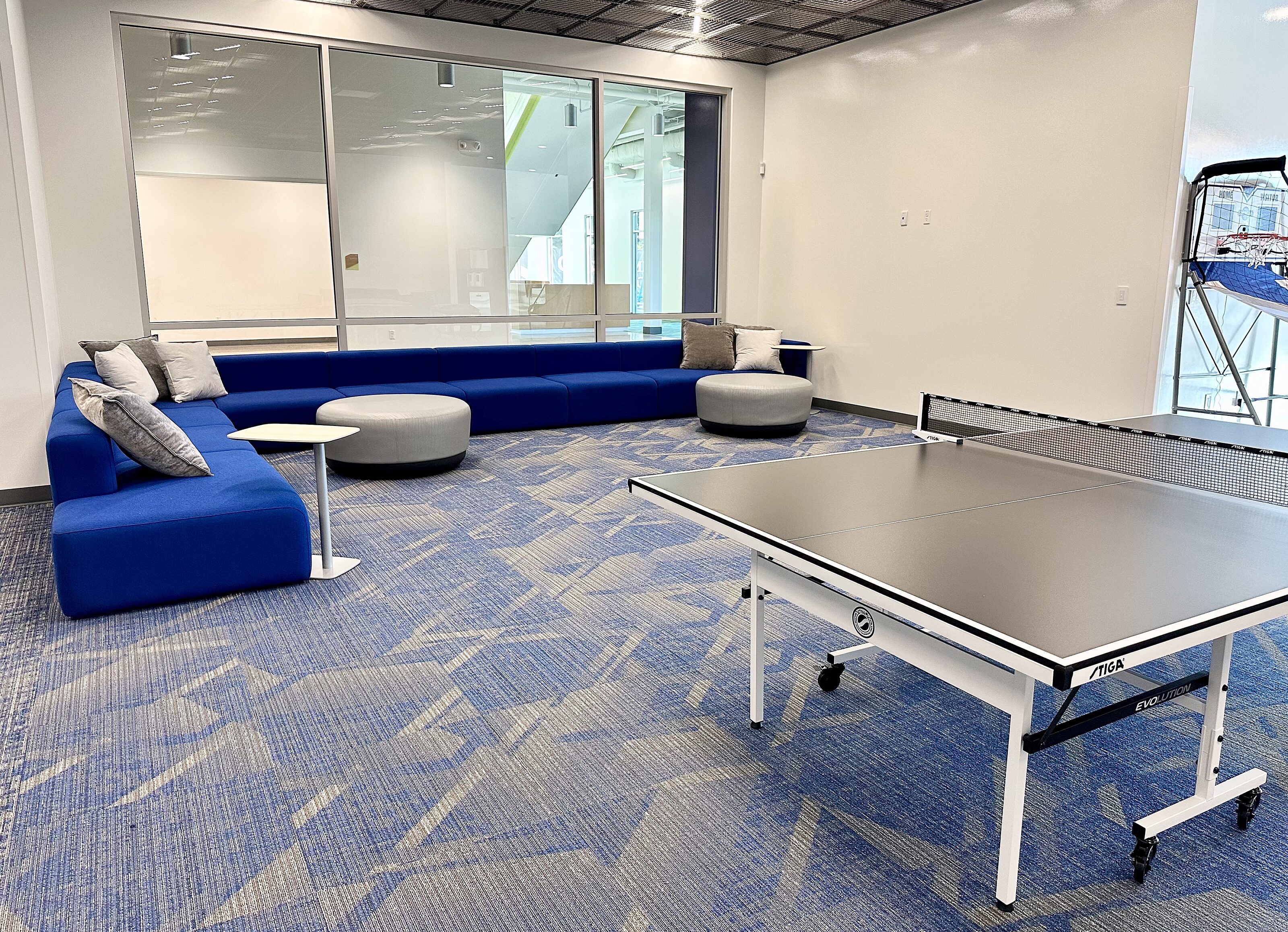 Relaxed space with a blue modular sofa, gray pillows, round ottomans, and a ping-pong table. Large windows enhance openness.