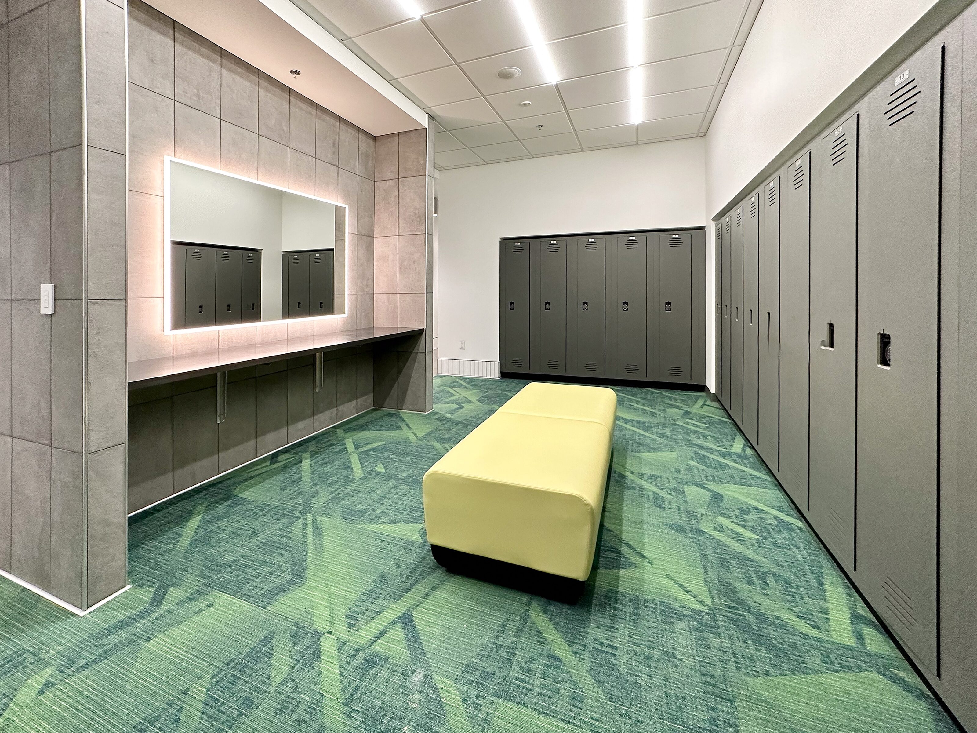 Sleek, modern locker room with green carpet, gray lockers, a vanity with an LED mirror, and a yellow cushioned bench.