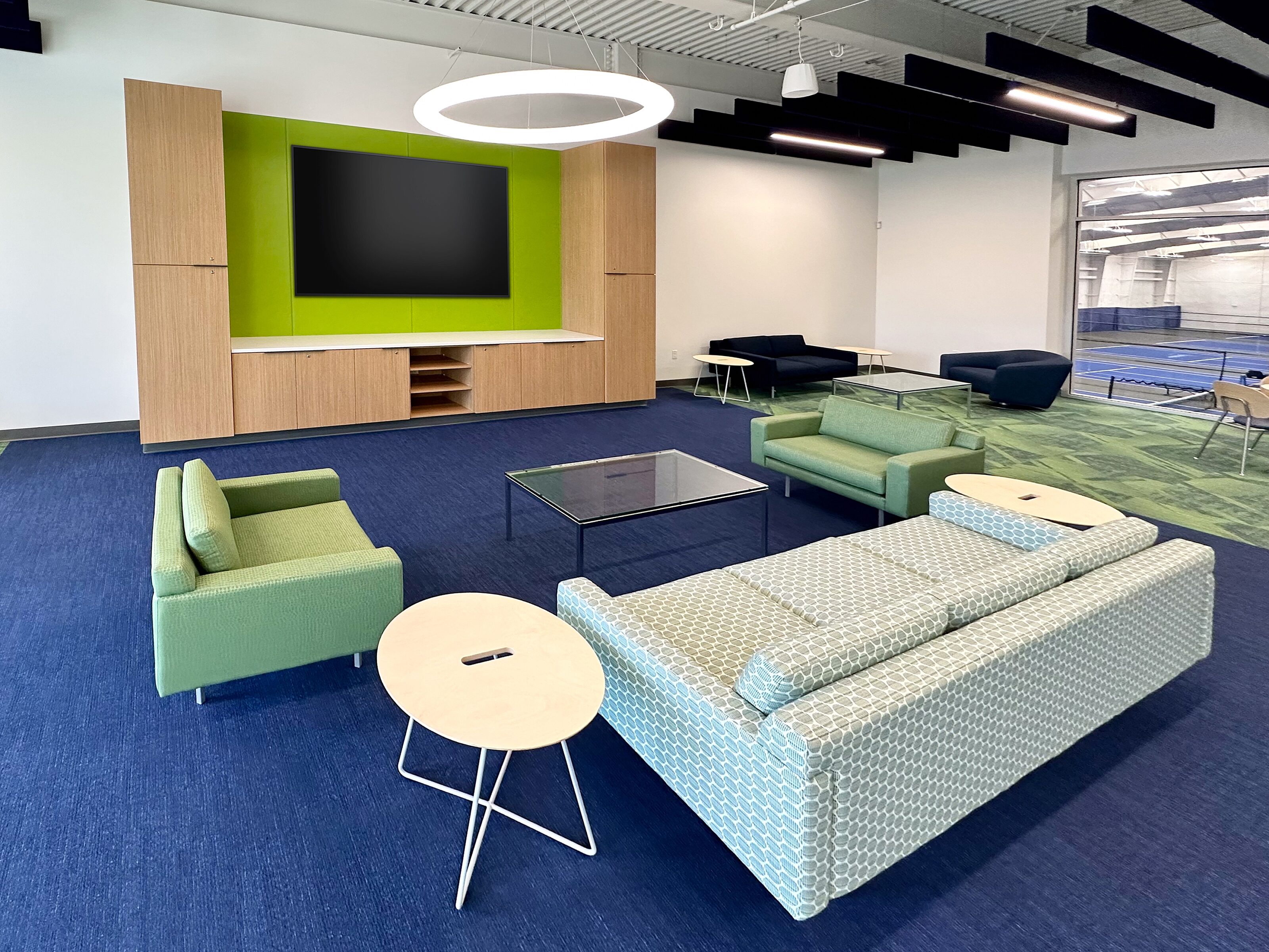 A stylish lounge with contemporary club chairs and sofa set in front of a media wall. Indoor tennis courts visible in the far distance.