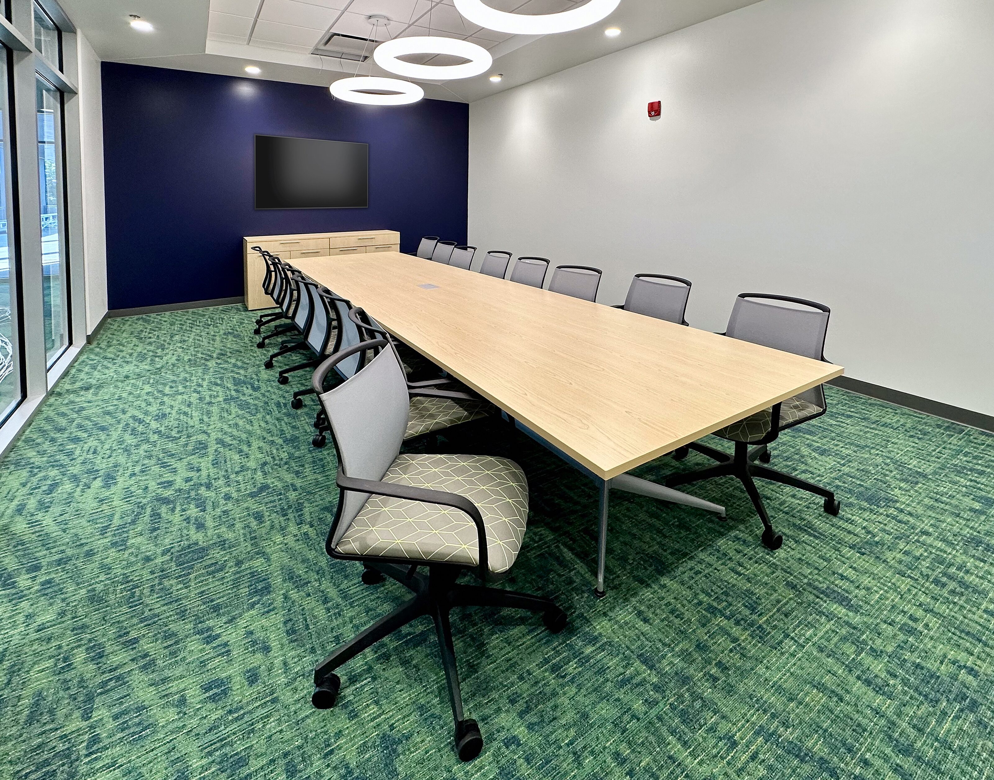 Professional setting with a long table, ergonomic chairs, a blue accent wall with a mounted screen, circular pendant lights, and large windows.