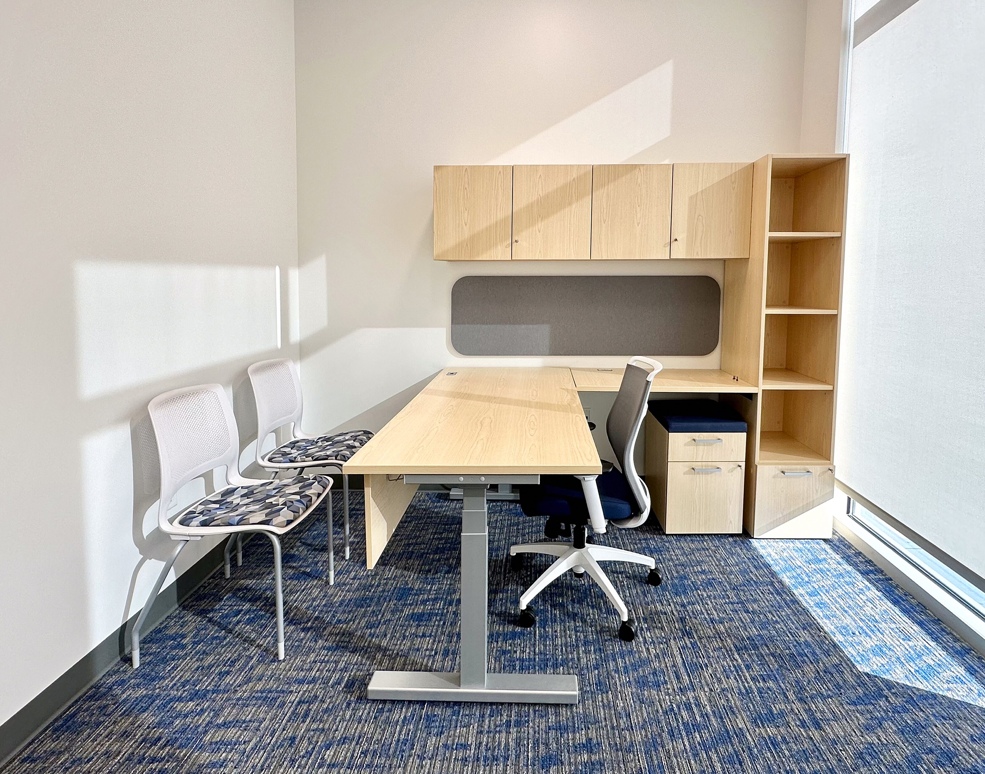 Sunlight illuminates a private office with L-shaped desk and ergonomic task chair. Overhead storage, open shelves, tackboard, and two guest chairs.
