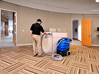 A uniformed worker stands next to a fabric-covered workstation wall. Using a rolling machine, they spply disinfectant to the furniture.