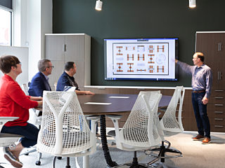 Four people meet at a tall conference table in a open space. Sunlight streams in from tall windows at left. Man points to presentation on screen.