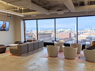 Office space on upper floor. Floor-to ceiling windows show downtown beyond. Cozy chairs & couches in foreground where person sits working. Concrete floor.