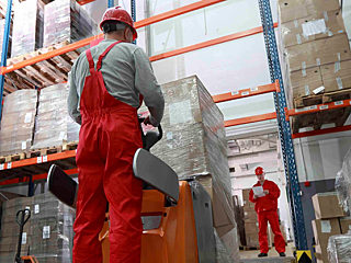 Low-angle view of a forklift operator in a bright warehouse. Tall shelving full of wrapped pallets, supervisor in distance with clipboard.