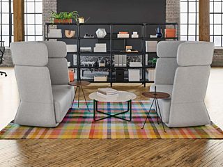 A loft space with exposed wooden beams, two high-back sofas, rug, and metal open shelf, surrounded by workstations and rolling task chairs