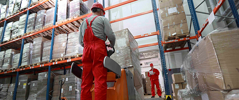 Low-angle view of a forklift operator in a bright warehouse. Tall shelving full of wrapped pallets, supervisor in distance with clipboard.