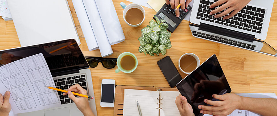Overhead view of people at a work table: hands, laptops, drawings, planners, pencils, coffee mugs