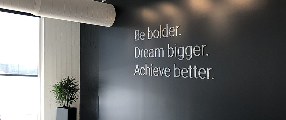 View of dark grey wall with inspirational brand message: Be bolder, dream bigger, achieve better. Windows to the left let in bright sunlight.