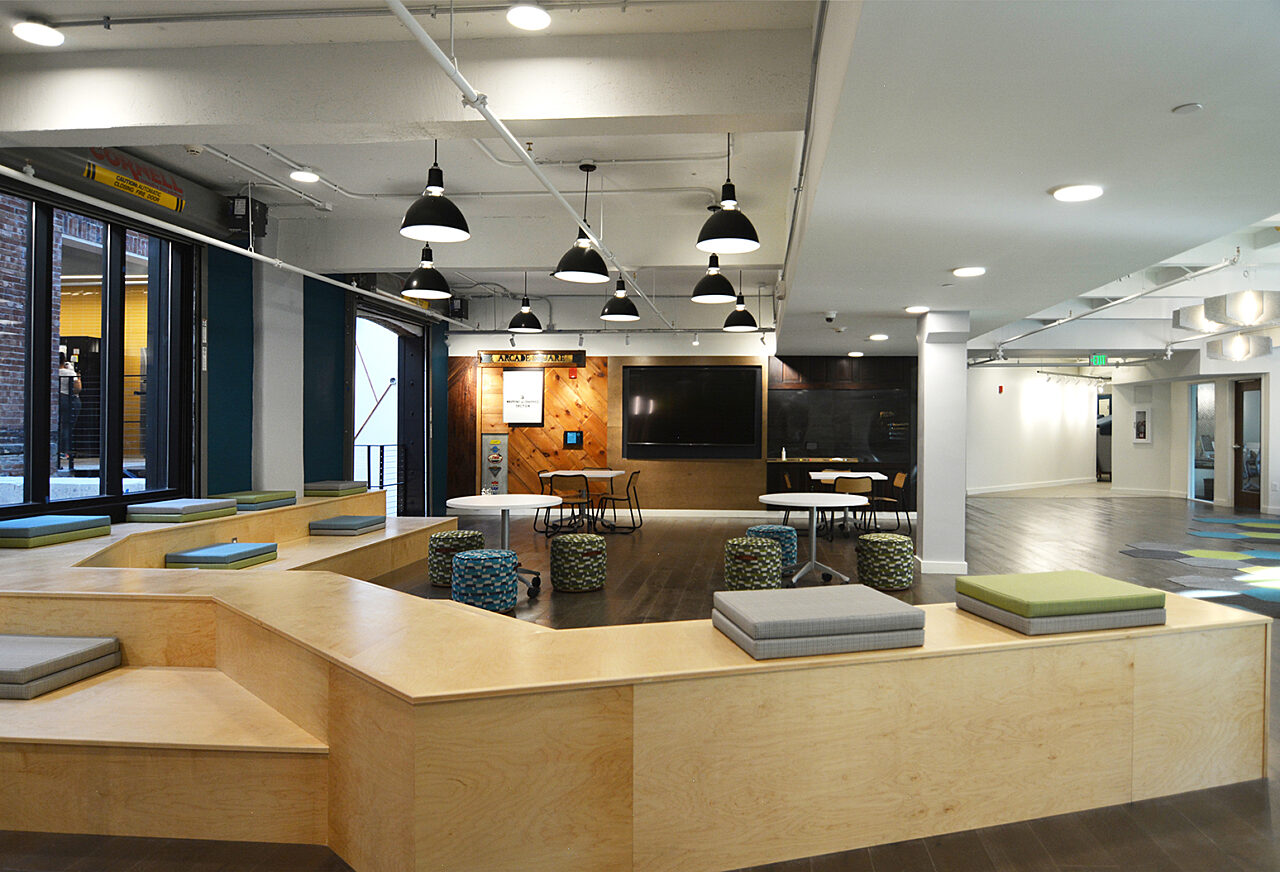 Modern office lounge with pendant lighting, wooden benches, and seating cushions around tables for gatherings.