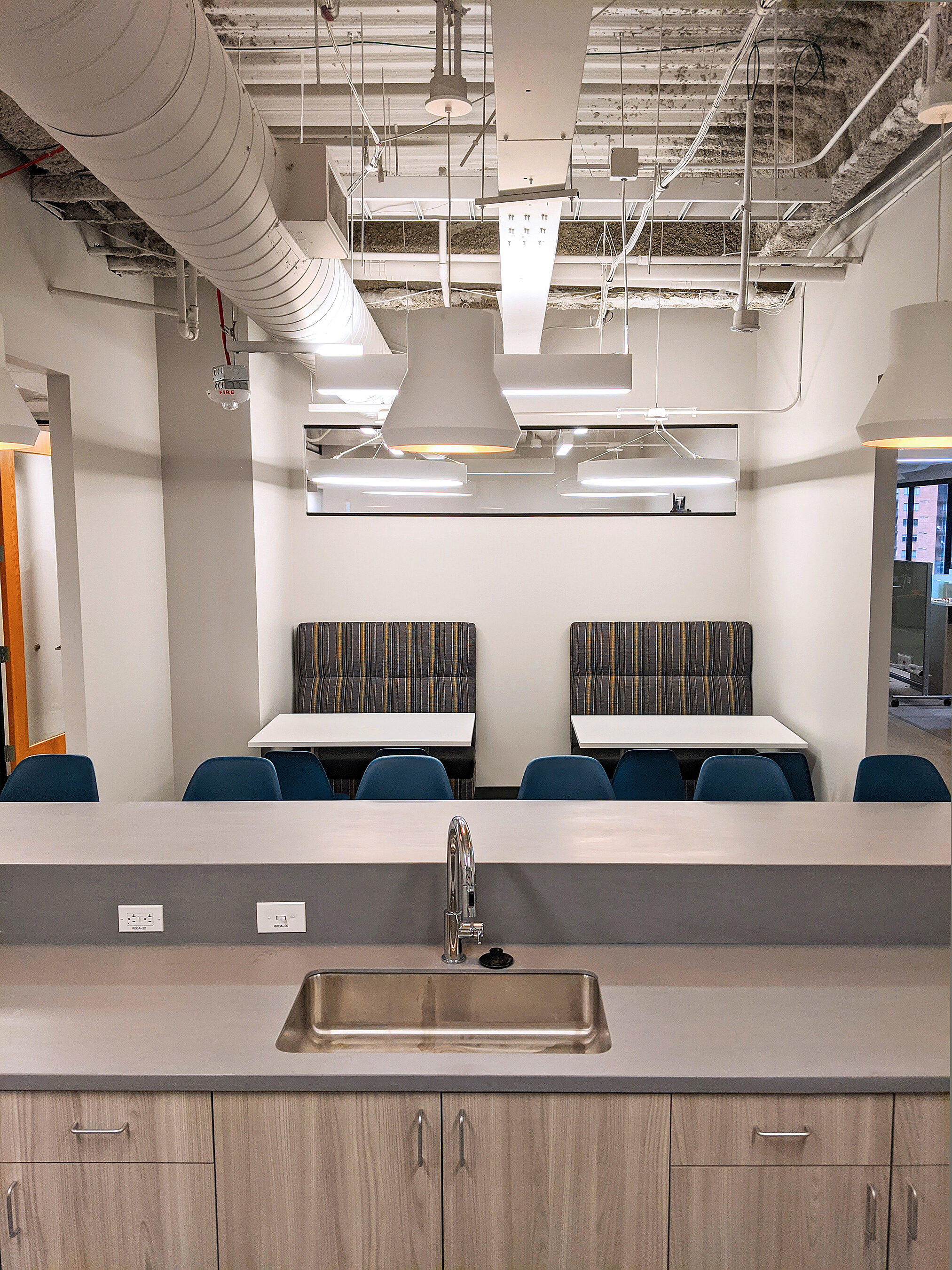Cafe with white walls & exposed beams & pipes. Sink in front with tall blue chairs. Booths in background feature dark patterned fabric & blue chairs