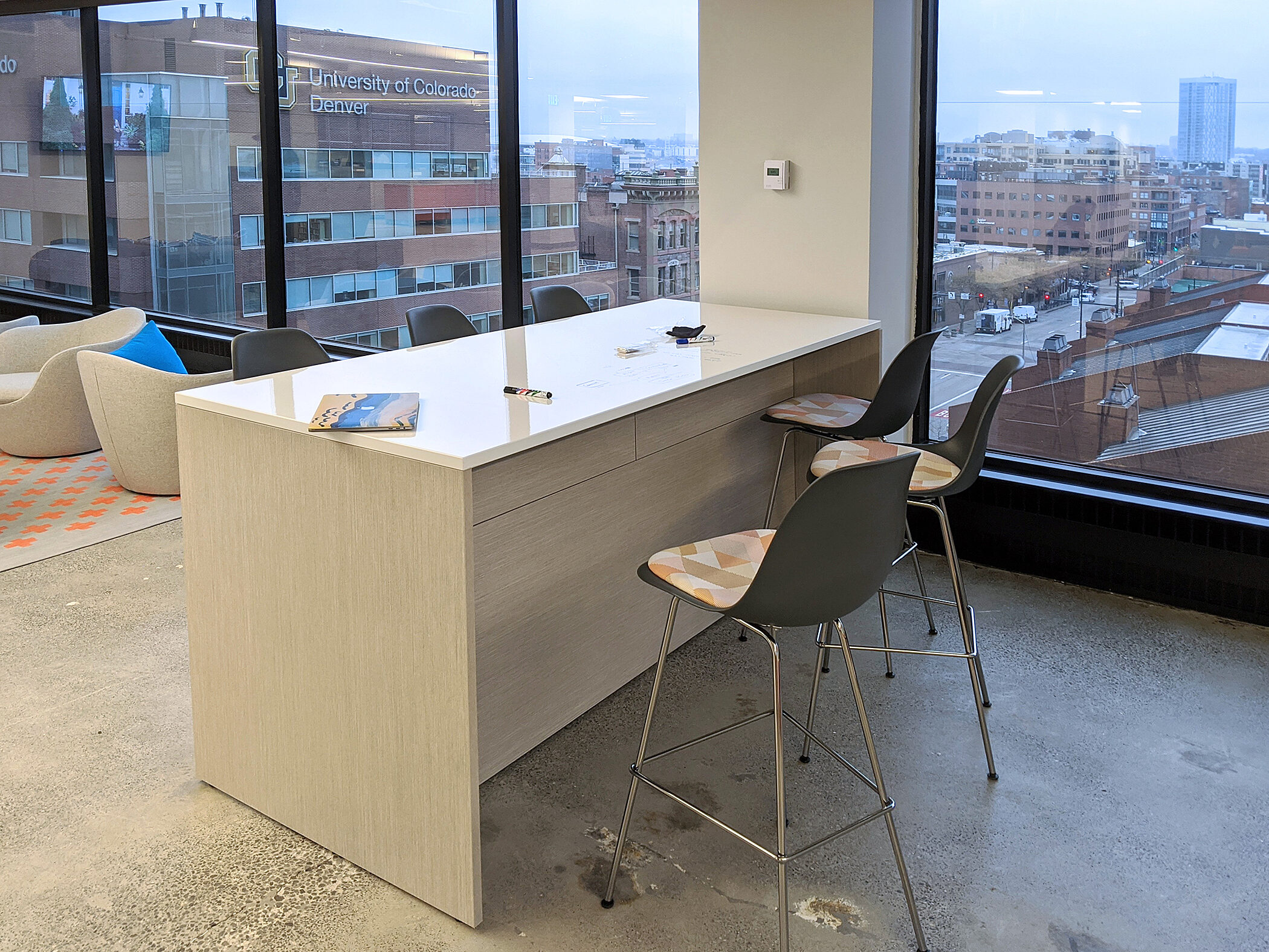 Tall, white table with waterfall sides as an island for casual meetings. Includes surface power and low-back stools. Large windows with view of city beyond