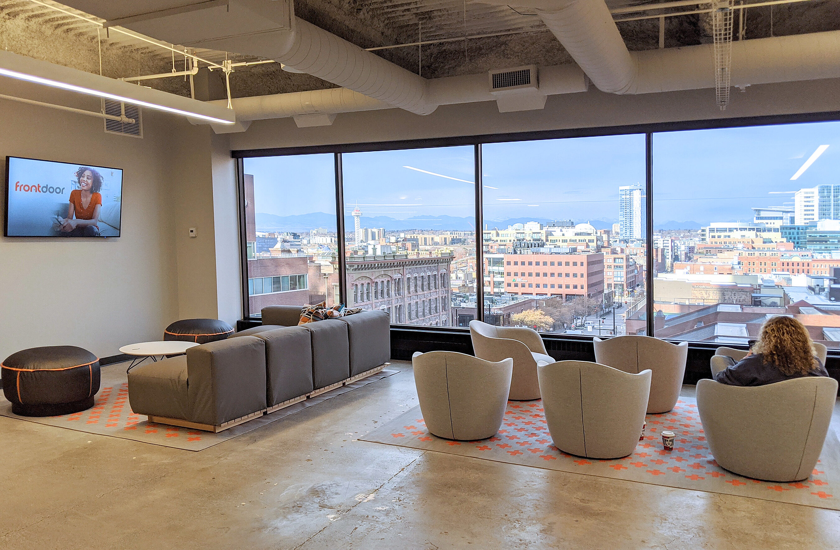 Office space on upper floor. Floor-to ceiling windows show downtown beyond. Cozy chairs & couches in foreground where person sits working. Concrete floor.