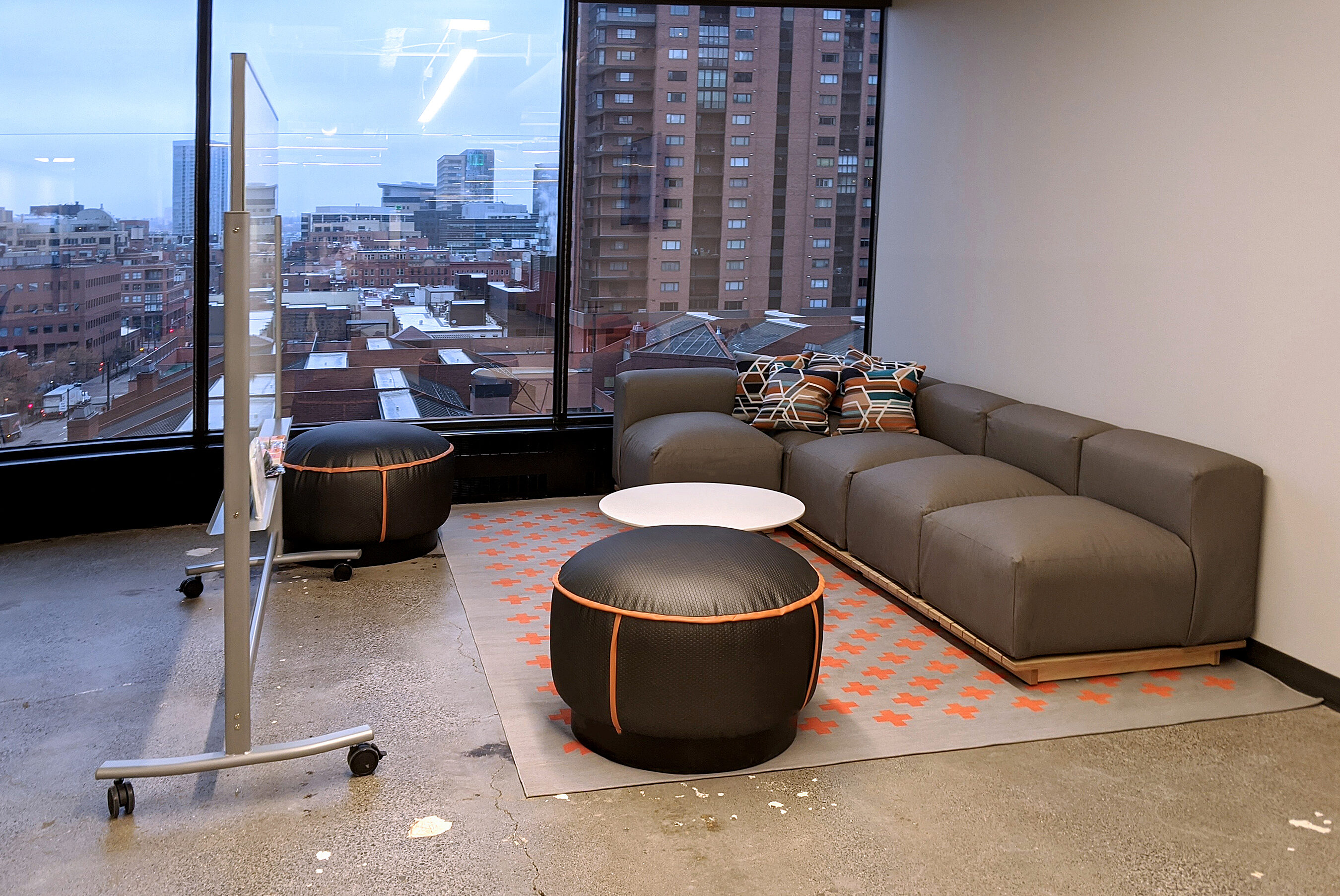 Open office collaborative area with low sofa and pouf-style seating and whiteboard. Large windows with view of city beyond.