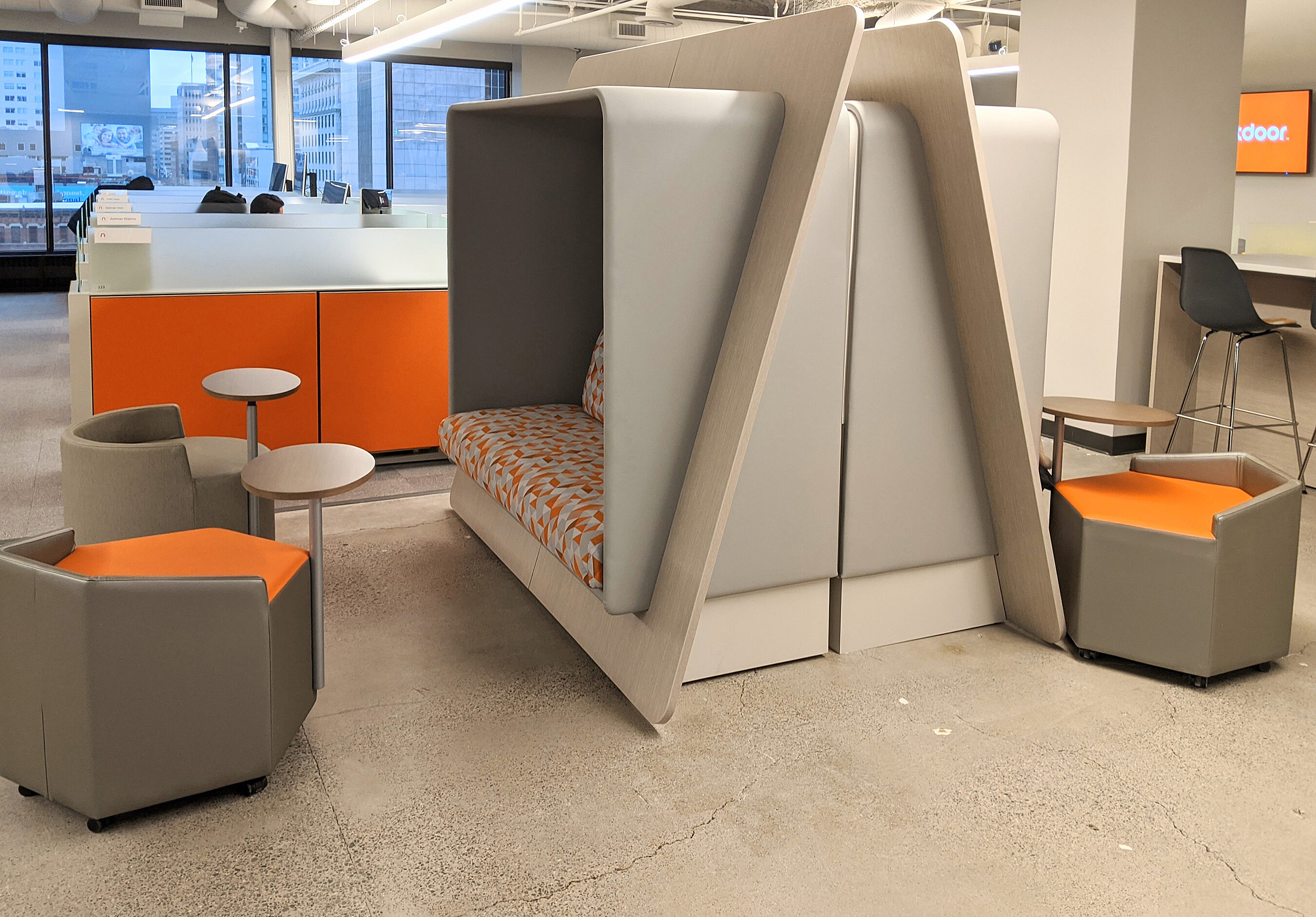 Cozy modern booth seating with octagonal soft chairs & tablet arms. Grey with orange accents. Workstations in background with orange exterior panels.