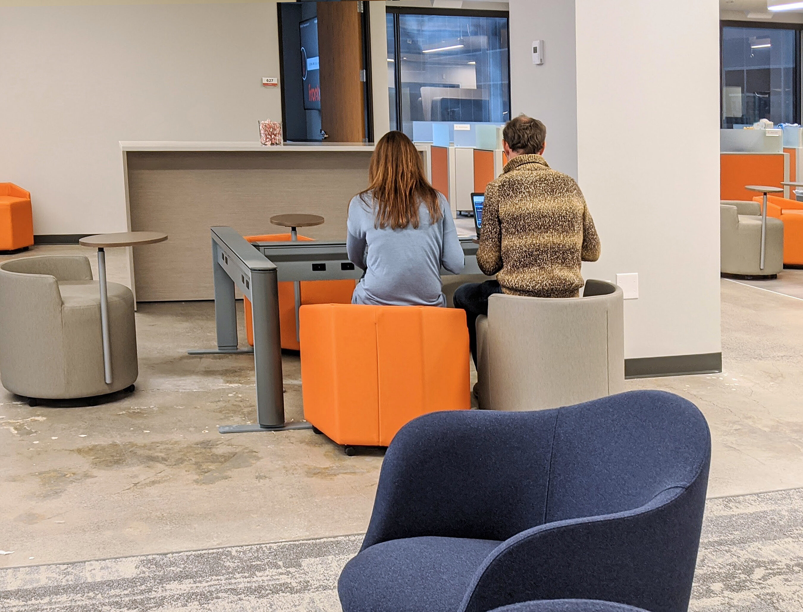 Office with two people in casual collab meeting at a powered notebook rail. Comfy soft seating in grey, orange, and blue. Concrete floor & white walls.