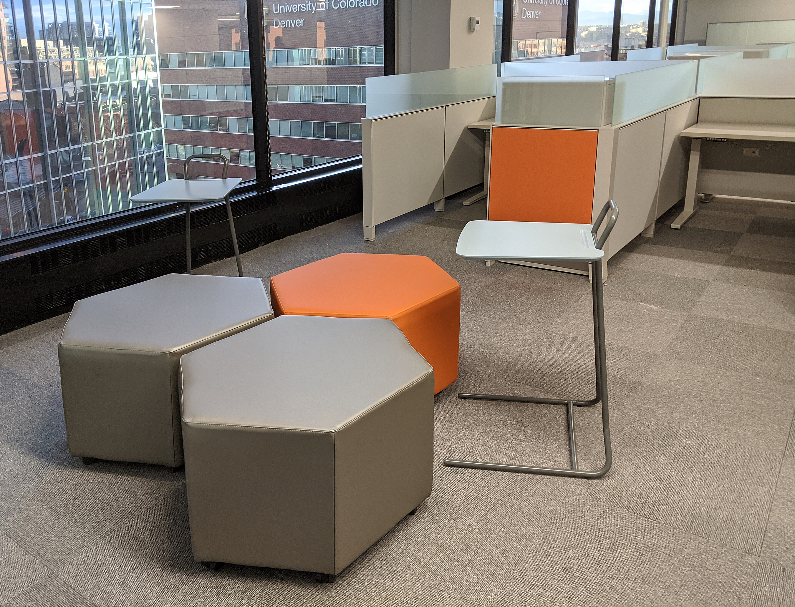 Large hexagonal ottomans function as tables or seating. Workspaces in near background and large windows offer view of city from high in the building.