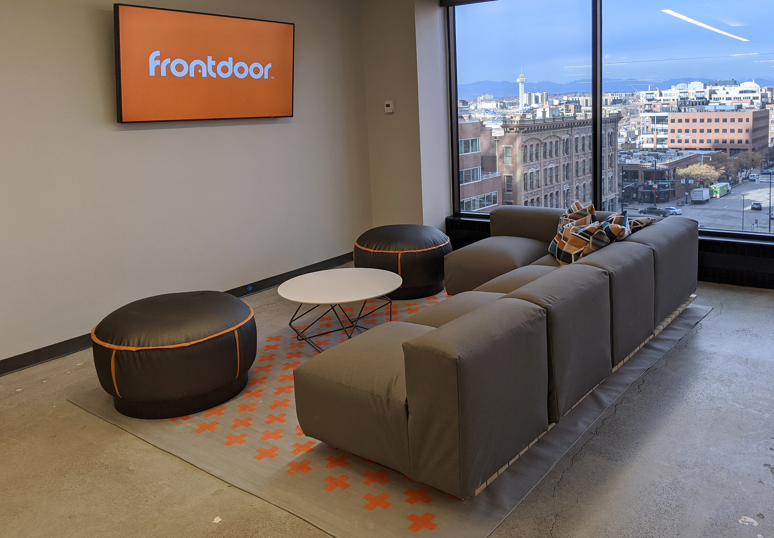 Office space in a tall building evident by floor-to ceiling windows that show city beyond. Low, soft couches & ottoman seats. Concrete floor & grey rug..