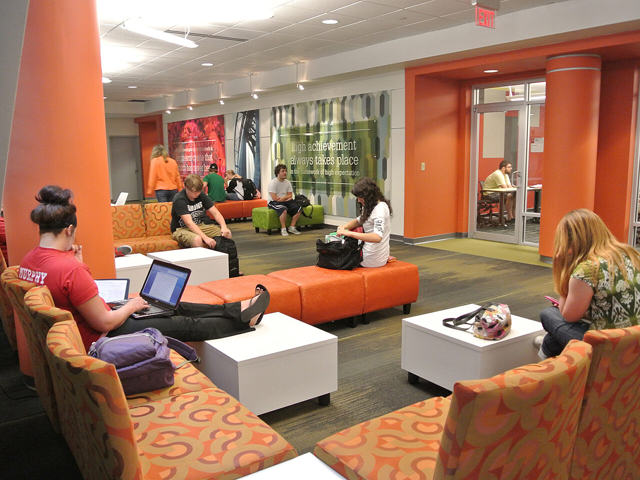 A group of individuals seated in an open space, surrounded by orange chairs and white tables, enjoying a casual gathering.