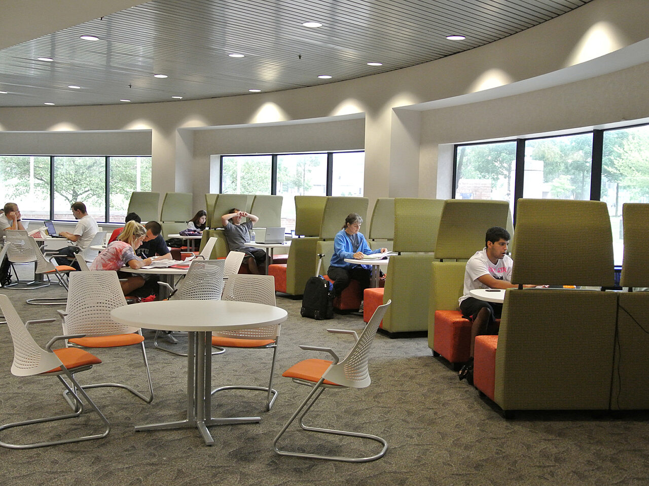 Individuals gathered at tables with green and orange seats in a bright, expansive room featuring large windows that enhance the atmosphere.